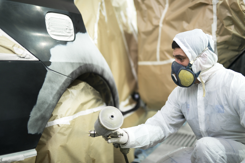 Man with protective clothes and mask painting automobile car bumper in repair shop.